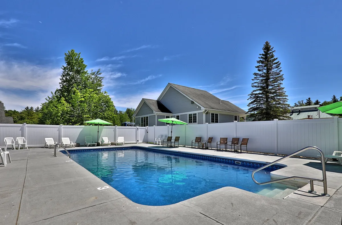 This image shows a vibrant outdoor swimming pool area bordered by a white fence and surrounded by lush greenery and trees. Green umbrellas shade the lounge chairs, inviting guests to relax poolside. The tranquil setting offers a glimpse of the comfort and leisure available on the property.