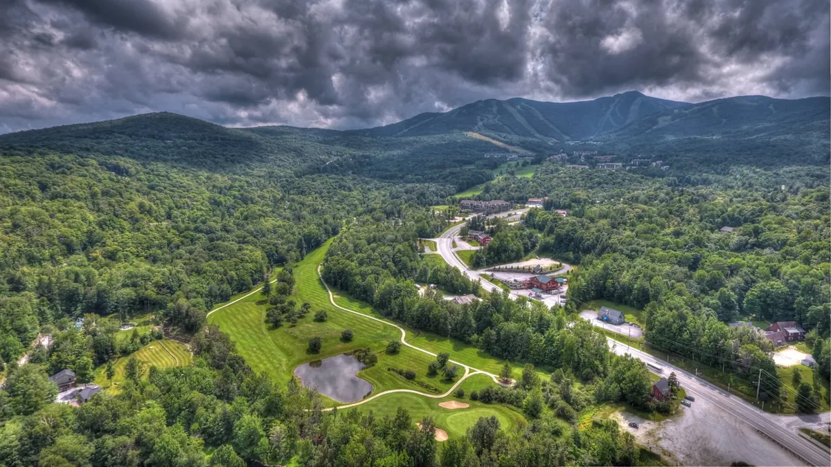 A sweeping exterior view of a scenic golf course nestled among rolling mountains and a reflective pond, surrounded by forested areas. The image exudes calm and showcases the natural beauty of the region.