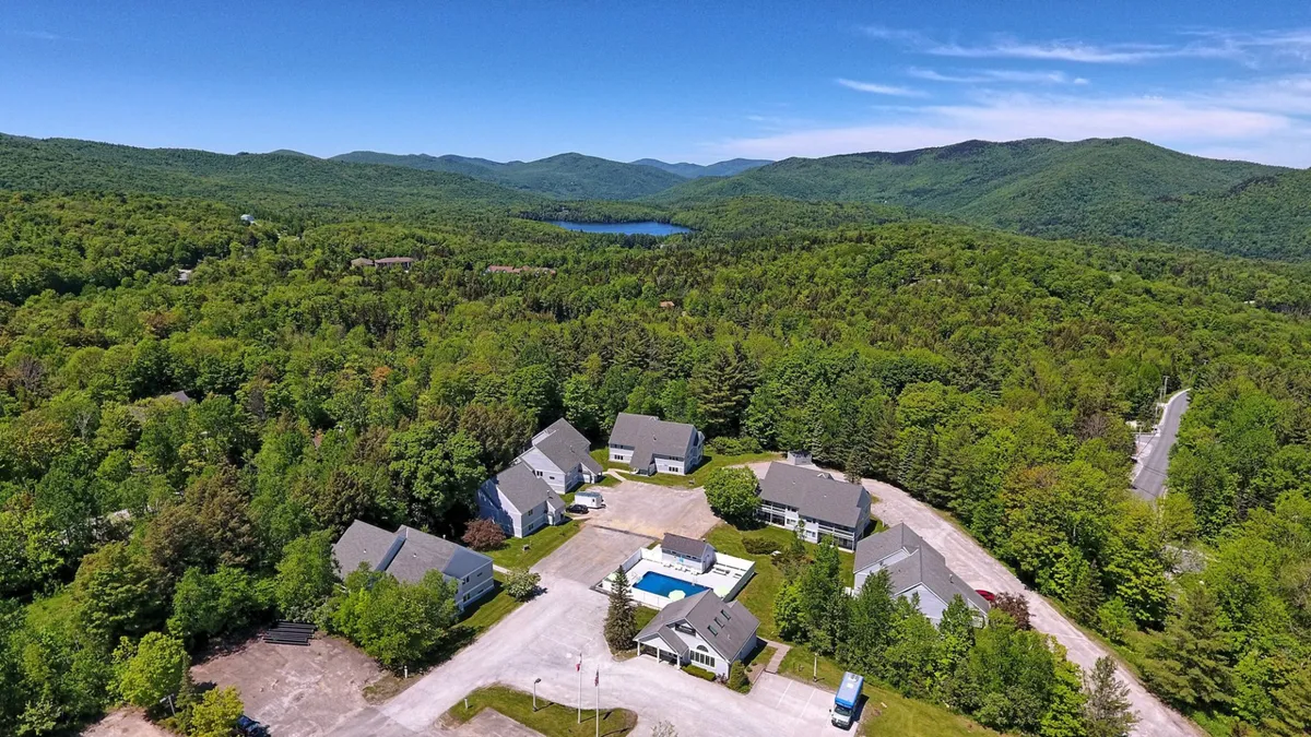 A sweeping exterior view capturing a mountain range in the background, a shimmering swimming pool, a cluster of buildings, and a distant lake. The image evokes a sense of connection to the outdoors, with communal spaces ideal for gatherings and relaxation.