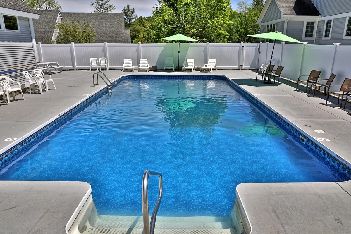 A bright poolside scene featuring an inviting swimming pool, lounge chairs, green umbrellas, and surrounding greenery. The setup reflects a lively, social, and relaxing summer environment.