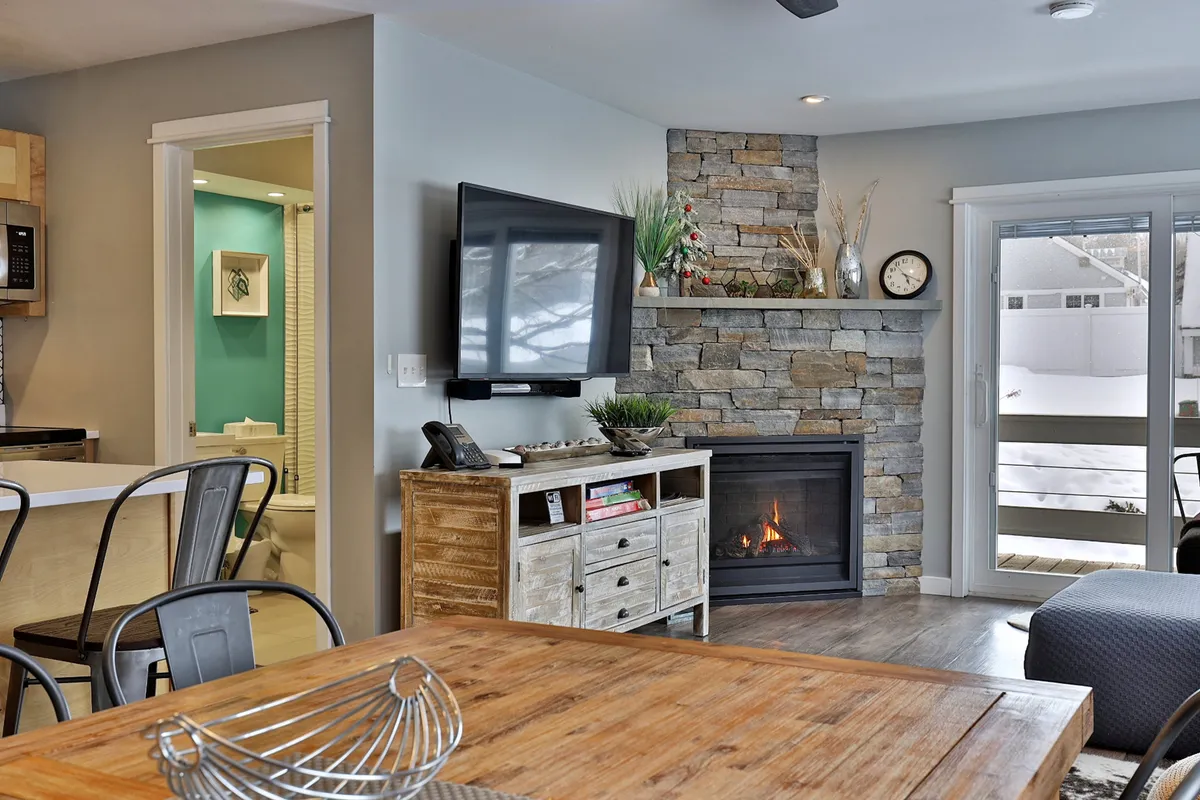 An inviting living-dining area featuring a fireplace, rustic wooden dining table, large TV, and access to a balcony. The arrangement suits both cozy evenings by the fire and lively meals, while the balcony doors hint at the connection between indoor comfort and outdoor possibilities.