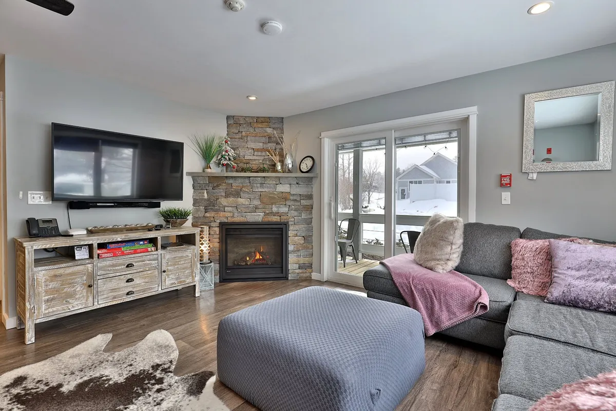 The image features a cozy, modern living area with a glowing fireplace as the centerpiece, a plush sofa facing a large television, a stylish coffee table, and a sliding glass door that fills the space with natural light and offers scenic views. The warm tones and inviting arrangement create a perfect spot for relaxing after a day on the slopes.