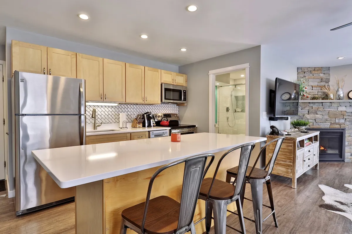 A modern kitchen with a central island, stainless steel appliances, and visible stone fireplace alongside dining chairs. The space feels open and conducive to both meal preparation and entertaining, with clear sightlines to the living area for easy socializing.