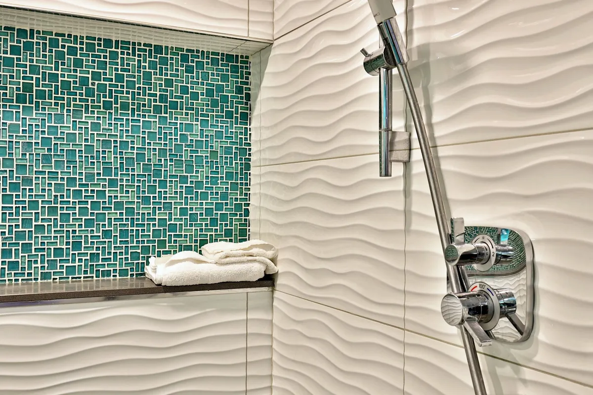 A serene bathroom interior featuring textured white tiles, a teal mosaic backsplash, modern shower fixtures, and neatly arranged towels on a shelf. The space feels spa-inspired, inviting relaxation and self-care.