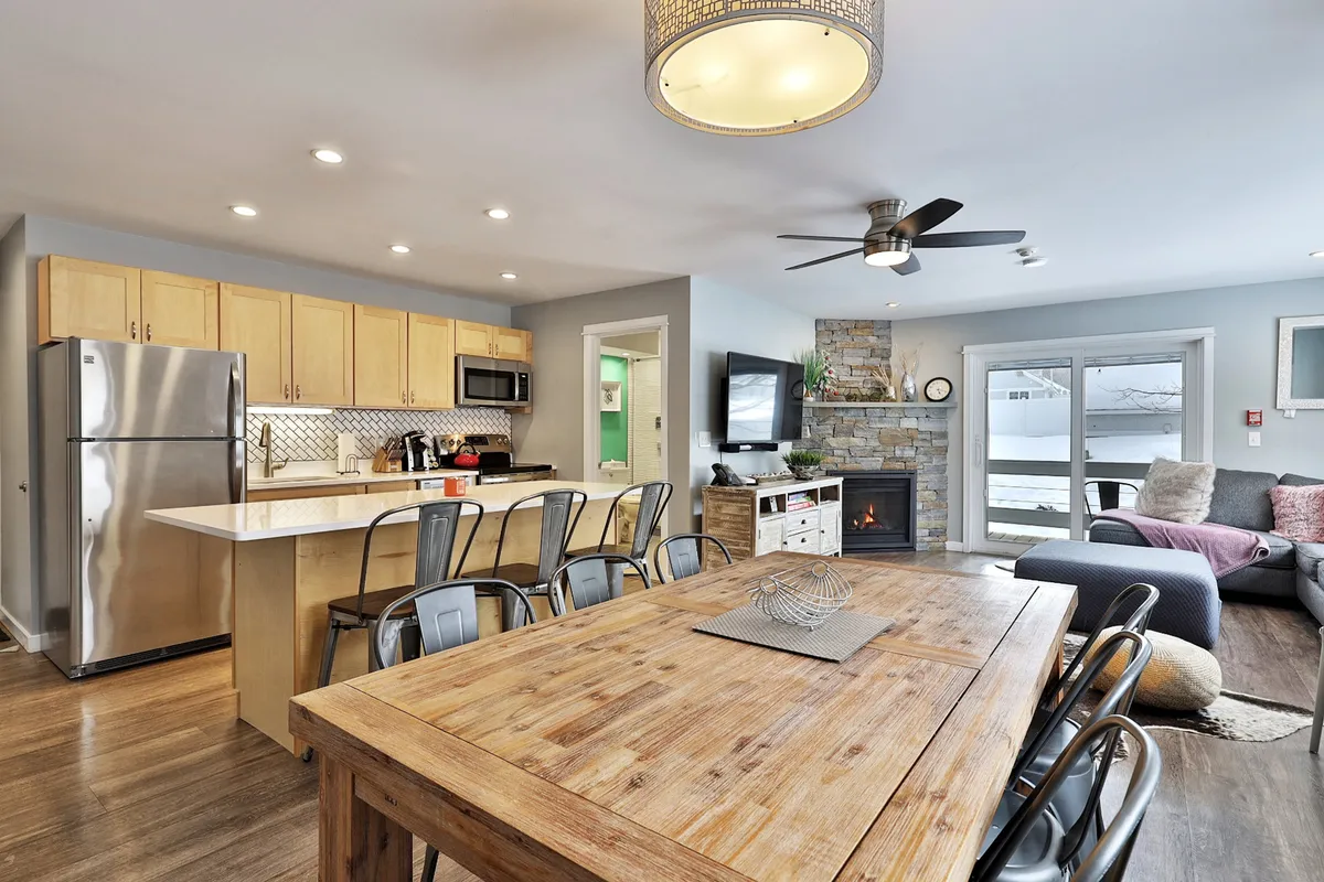 A cozy, open-plan interior featuring a dining table, kitchen island, stone fireplace, and a comfortable sofa. The living area is warmly lit with natural light streaming through the windows, and the arrangement invites both relaxation and social gatherings. The fireplace and sofa set the scene for movie nights, while the dining and kitchen areas connect seamlessly for group meals and conversation.