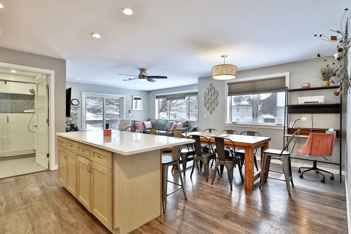 An airy interior featuring a kitchen island and dining table, both bathed in natural light streaming through large windows. The decorative wall art adds personality, while the open layout lends itself to both productivity and comfort. The space is depicted as comfortable and flexible, suitable for working from home or enjoying casual gatherings.