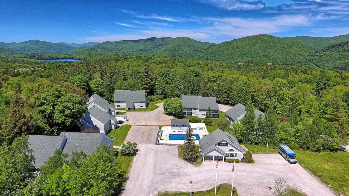 This exterior image showcases a heated outdoor swimming pool set against a striking mountain landscape, with residential buildings and lush greenery surrounding the scene. Lounge chairs and the tranquil setting evoke leisure and summer relaxation.