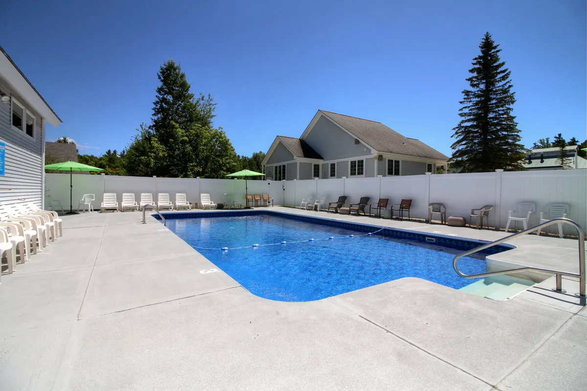 A broad view of the outdoor pool area with lounge chairs, poolside umbrellas, and buildings in the background. The surrounding greenery enhances the peaceful, resort-like feel, offering plenty of space for relaxation and socializing.