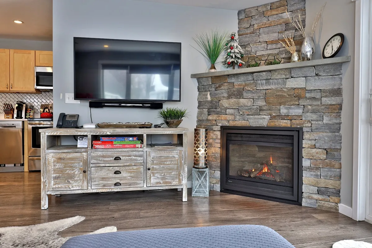 This interior image features a welcoming gas fireplace with a stone surround, complemented by seasonal decor, a cozy seating area, and a television. The inviting space is perfect for warming up and unwinding after a day outdoors in the snow.