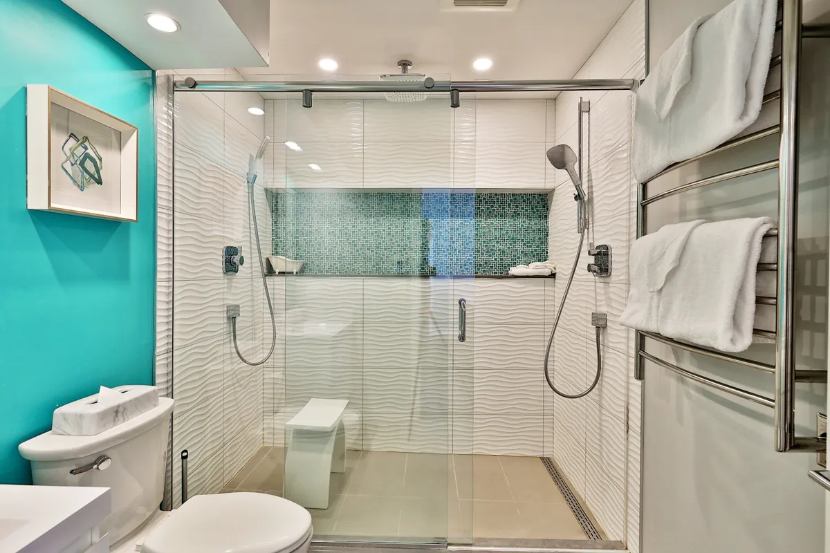 An inviting bathroom with a glass shower enclosure, textured wall tiles, a teal accent wall, mosaic backsplash, and a towel rack with plush white towels. The ambiance is both fresh and serene, ideal for invigorating morning routines or relaxing evening baths.